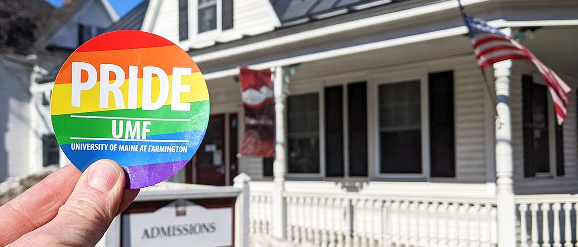 A University of Maine at Farmington LGBTQ+ Pride sticker held in front of a UMF campus building in the background.