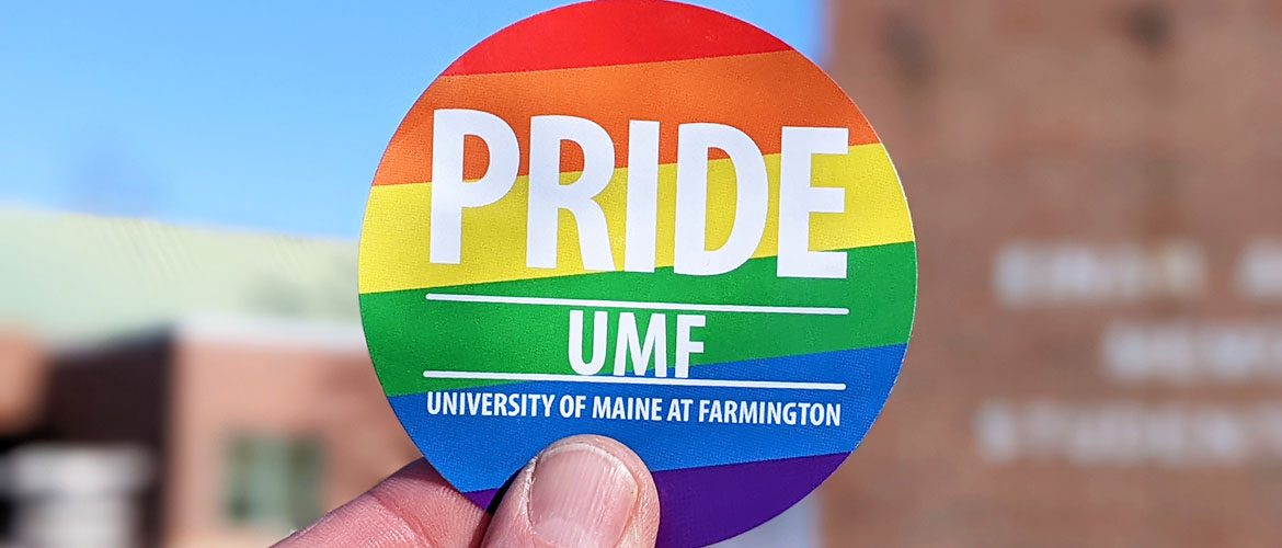 A University of Maine at Farmington LGBTQ+ Pride sticker held in front of a UMF campus building in the background.