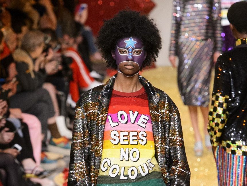 image of a woman walking on a runway, depicted from the waist up. She is wearing a horizontally striped rainbow shirt with the words "love sees no colour" in white lettering under a sequined, brown, plaid suit jacket