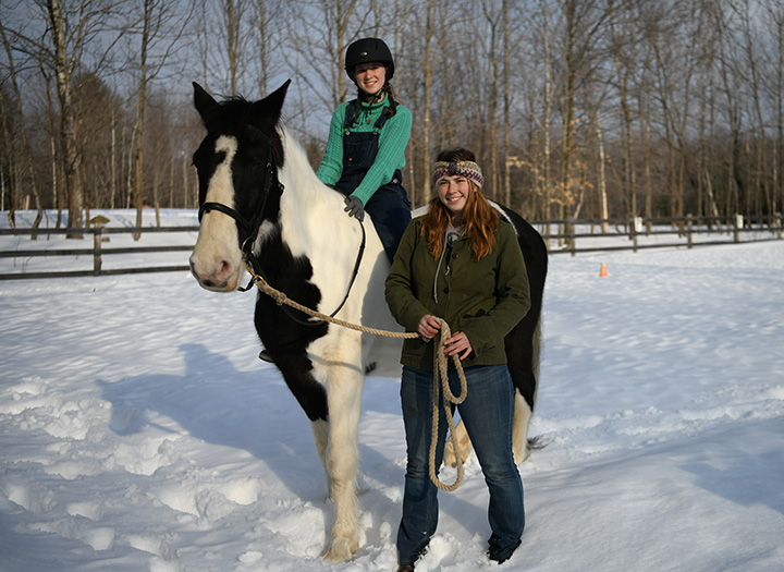 Jessica Cloutier (standing), a recent graduate from Corinna in the University of Maine at Farmington’s Rehabilitation Services program and coach for the UMF Equestrian Club, helps Tori Oliveira, UMF student and Equestrian Club member, as she rides Big Mike for some winter exercise.
